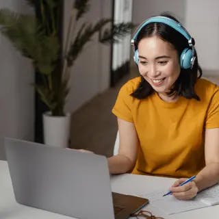 Picture of woman wearing ochre coloured t-shirt wearing light blue headphones, sitting at a desk and smiling at laptop screen